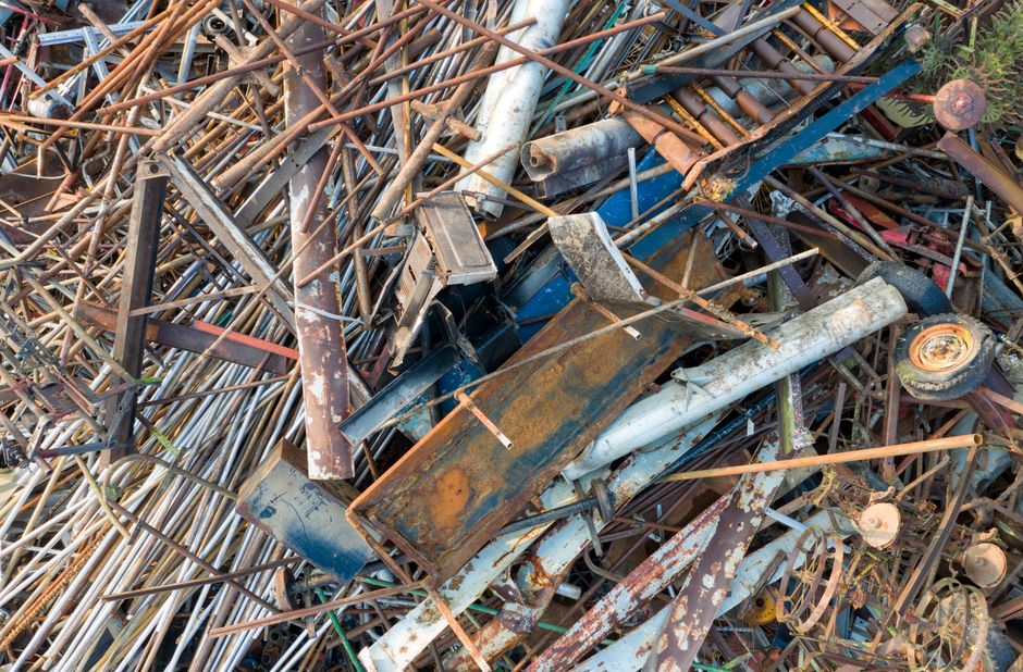 A large pile of discarded metal scrap on a flat outdoor surface, consisting of rusted steel and aluminum rods, beams, pipes, and mechanical parts with varying textures from smooth to corroded. The met